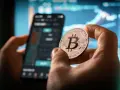 Dłoń trzyma monetę z symbolem Bitcoina, na tle wykresów i ekranu telefonu. Może to być nawiązanie do historii Jamesa Howelsa i jego utraconych bitcoinów.