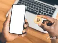 Dłoń trzyma smartfon z pustym ekranem i monetę bitcoin. Możliwe, że to początek przygody z kopanie kryptowalut na telefonie.