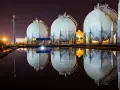 Duże, białe zbiorniki magazynujące gaz ziemny (natgas) odbijają się w wodzie. W tle nocne oświetlenie.