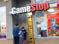 Przed sklepem GameStop akcje, ludzie w maskach rozmawiają. W witrynie gry i reklama "DOMINATE FROM DAY 1".