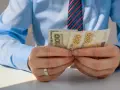 Mężczyzna w niebieskiej koszuli i krawacie liczy banknoty 100 i 200 zł. Może to być związane z rozliczeniem zus preferencyjny.