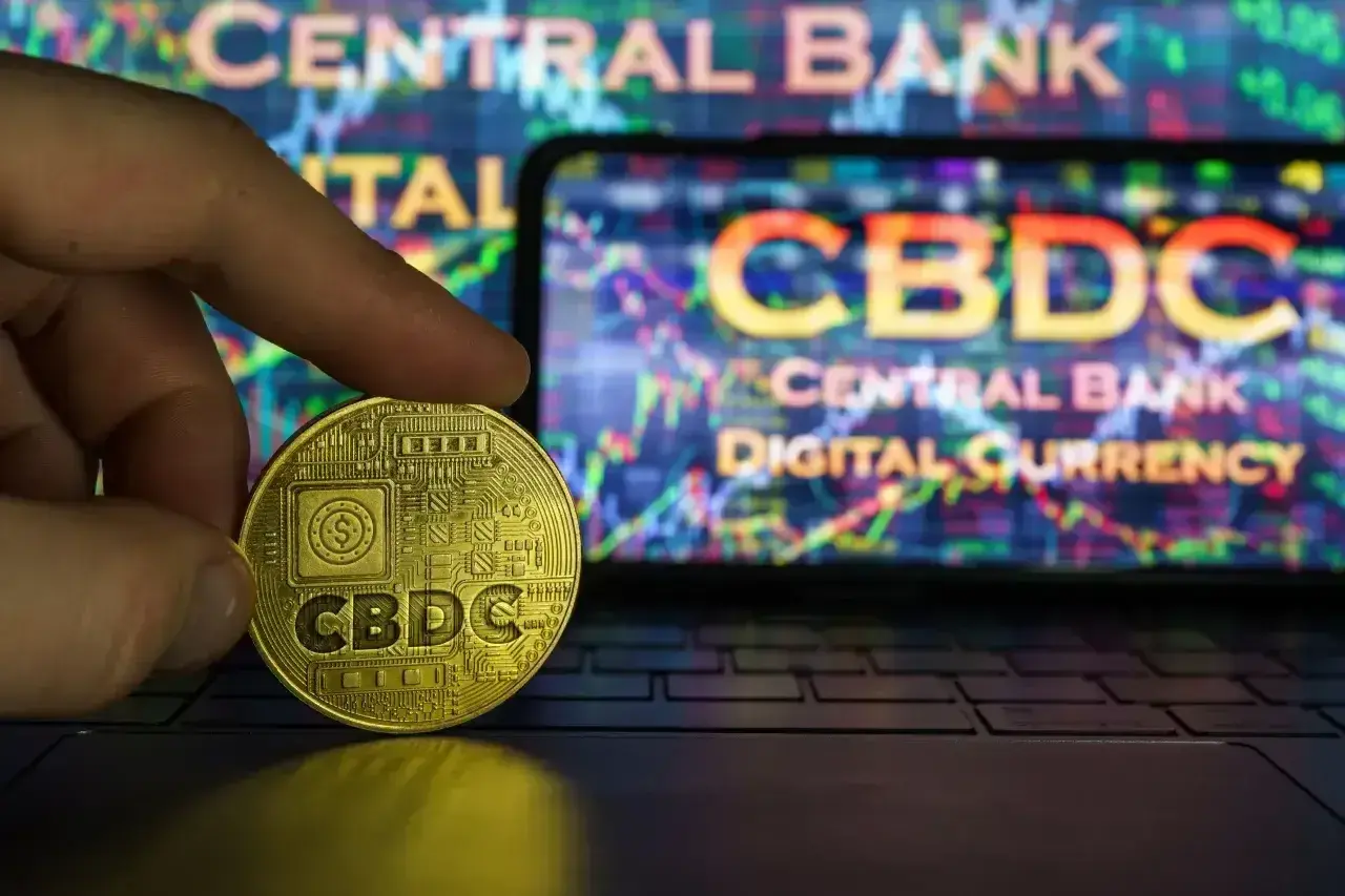 Złoty żeton z napisem CBDC w dłoni, na tle ekranu z wykresem i napisem "CENTRAL BANK DIGITAL CURRENCY". Potencjalne zagrożenia CBDC.
