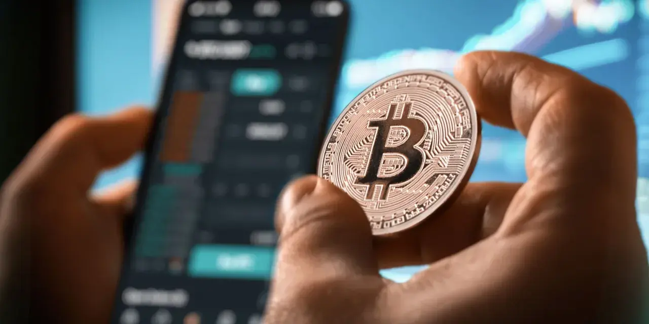 Dłoń trzyma monetę z symbolem Bitcoina, na tle wykresów i ekranu telefonu. Może to być nawiązanie do historii Jamesa Howelsa i jego utraconych bitcoinów.