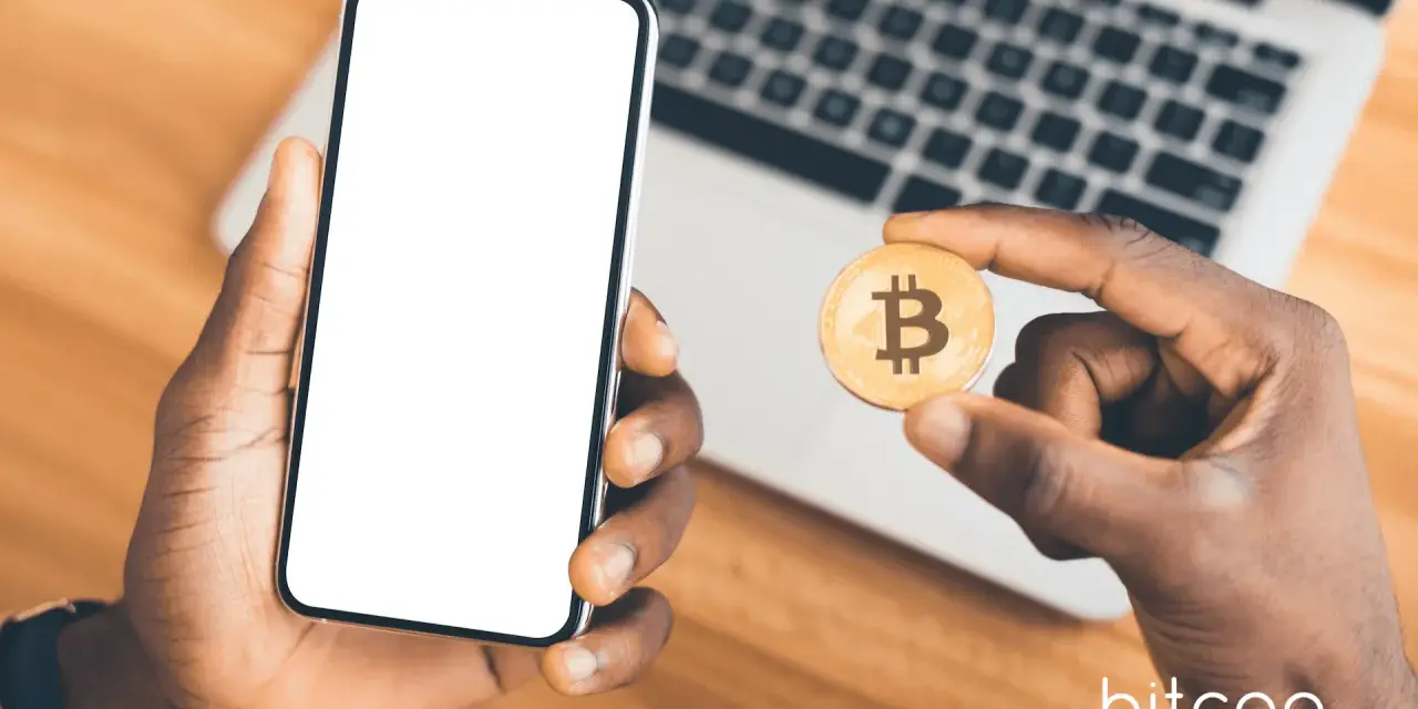 Dłoń trzyma smartfon z pustym ekranem i monetę bitcoin. Możliwe, że to początek przygody z kopanie kryptowalut na telefonie.