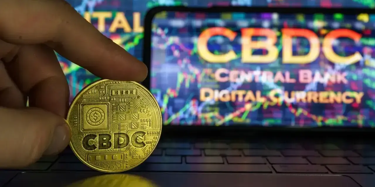 Złoty żeton z napisem CBDC w dłoni, na tle ekranu z wykresem i napisem "CENTRAL BANK DIGITAL CURRENCY". Potencjalne zagrożenia CBDC.