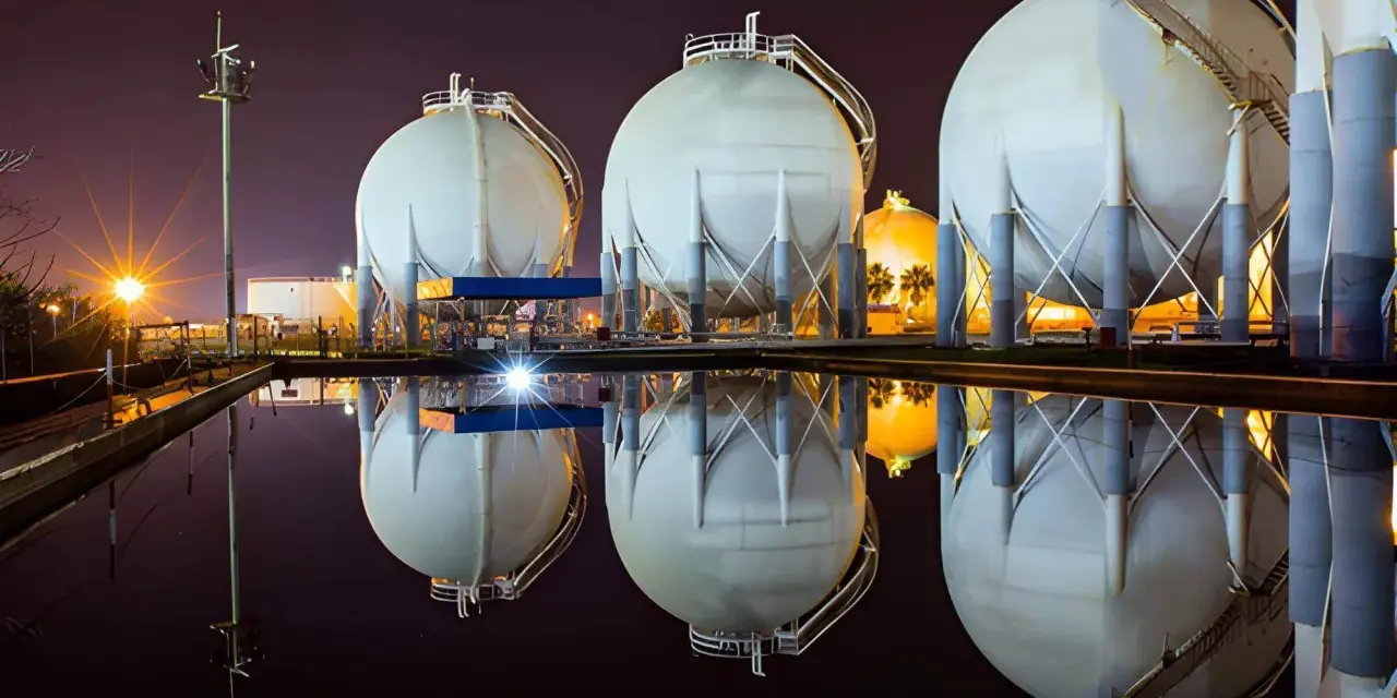 Duże, białe zbiorniki magazynujące gaz ziemny (natgas) odbijają się w wodzie. W tle nocne oświetlenie.