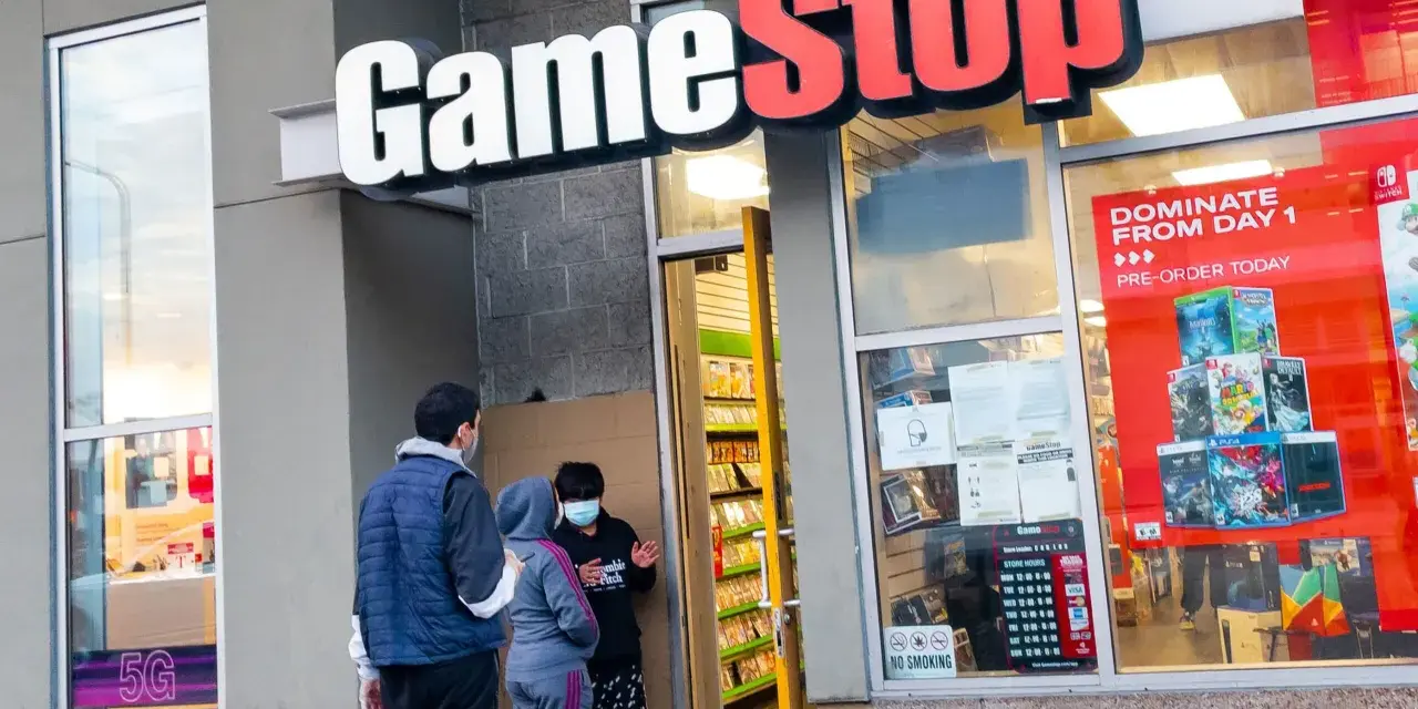 Przed sklepem GameStop akcje, ludzie w maskach rozmawiają. W witrynie gry i reklama "DOMINATE FROM DAY 1".