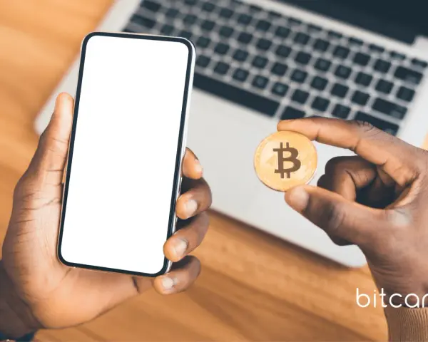 Dłoń trzyma smartfon z pustym ekranem i monetę bitcoin. Możliwe, że to początek przygody z kopanie kryptowalut na telefonie.