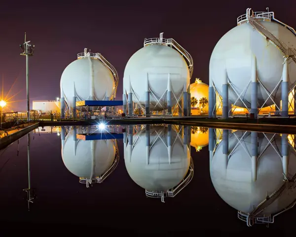 Duże, białe zbiorniki magazynujące gaz ziemny (natgas) odbijają się w wodzie. W tle nocne oświetlenie.
