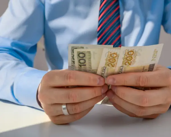 Mężczyzna w niebieskiej koszuli i krawacie liczy banknoty 100 i 200 zł. Może to być związane z rozliczeniem zus preferencyjny.