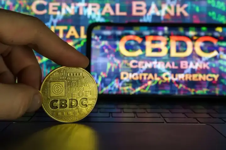 Złoty żeton z napisem CBDC w dłoni, na tle ekranu z wykresem i napisem "CENTRAL BANK DIGITAL CURRENCY". Potencjalne zagrożenia CBDC.