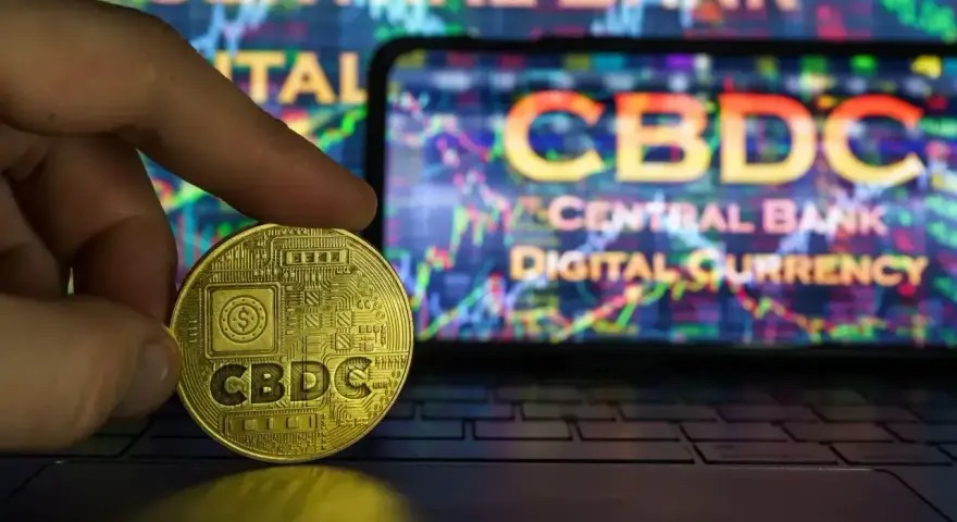 Złoty żeton z napisem CBDC w dłoni, na tle ekranu z wykresem i napisem "CENTRAL BANK DIGITAL CURRENCY". Potencjalne zagrożenia CBDC.