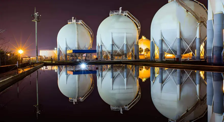 Duże, białe zbiorniki magazynujące gaz ziemny (natgas) odbijają się w wodzie. W tle nocne oświetlenie.