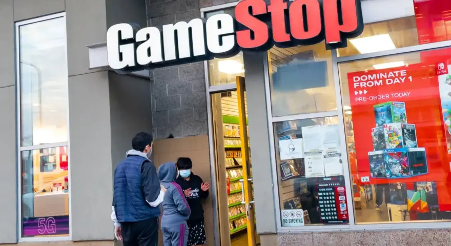 Przed sklepem GameStop akcje, ludzie w maskach rozmawiają. W witrynie gry i reklama "DOMINATE FROM DAY 1".