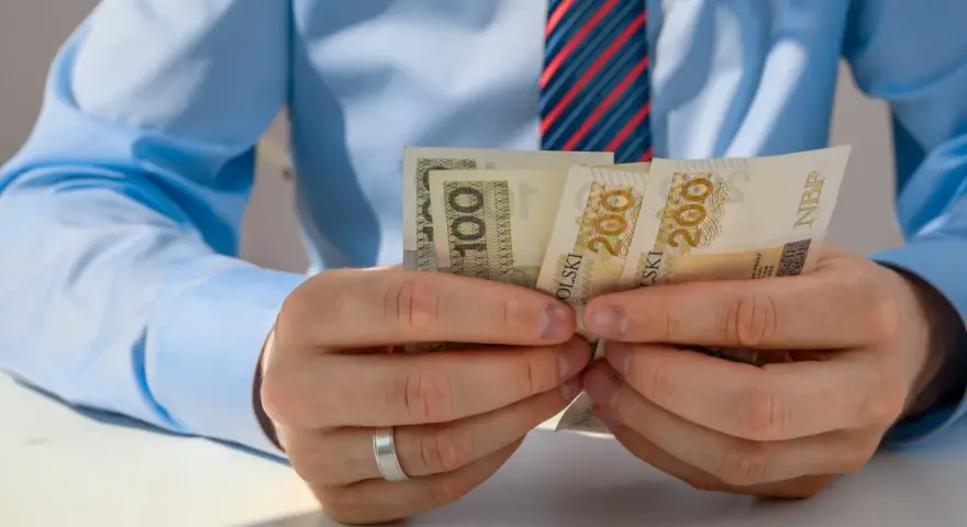 Mężczyzna w niebieskiej koszuli i krawacie liczy banknoty 100 i 200 zł. Może to być związane z rozliczeniem zus preferencyjny.